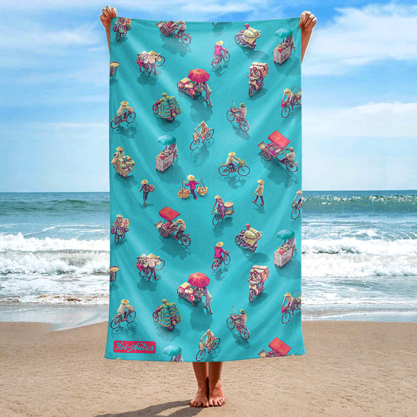Front view of a large blue beach towel, held up at the beach, with a pattern of people on bicycles and motorcycles, featuring Hoi-Antics brand logo.