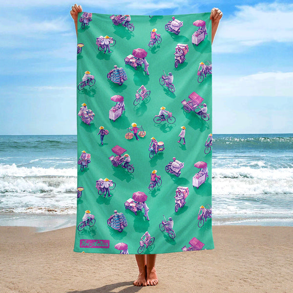 Front view of a large green beach towel, held up at the beach, with a pattern of people on bicycles and motorcycles, featuring Hoi-Antics brand logo.