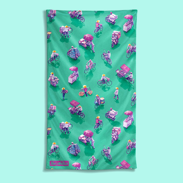 Front view of a large green beach towel with a pattern of people on bicycles and motorcycles, featuring Hoi-Antics brand logo.