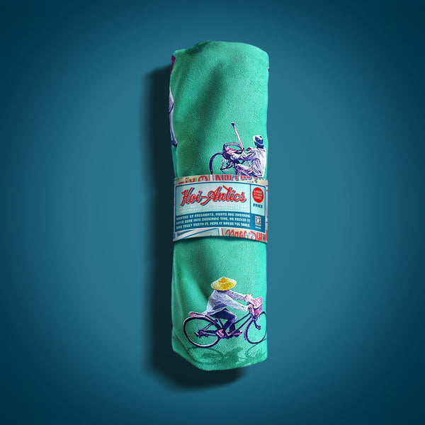 Front view of a rolled up large green beach towel with a pattern of people on bicycles and motorcycles, featuring Hoi-Antics brand wrapper.