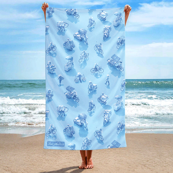 Front view of a large light blue beach towel, held up at the beach, with a pattern of people on bicycles and motorcycles, featuring Hoi-Antics brand logo.