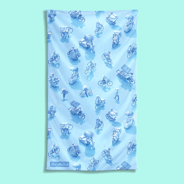 Front view of a large light blue beach towel with a pattern of people on bicycles and motorcycles, featuring Hoi-Antics brand logo.