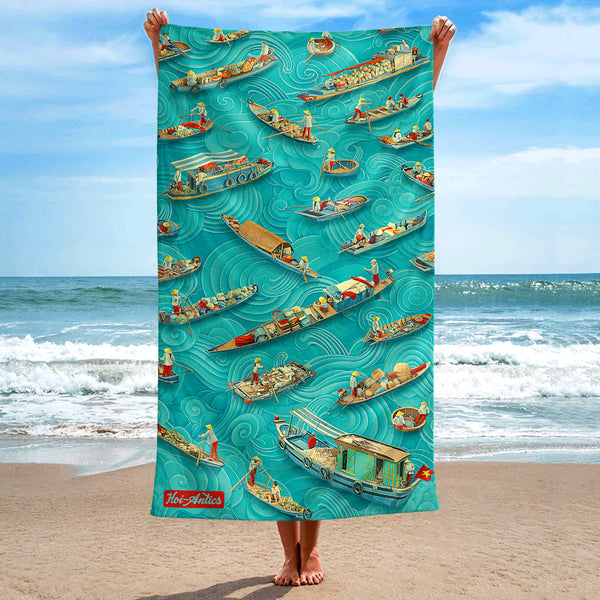 Front view of a large Mekong beach towel, held up at the beach, with a pattern of boats and people floating on asian inspired waves, featuring Hoi-Antics brand logo.
