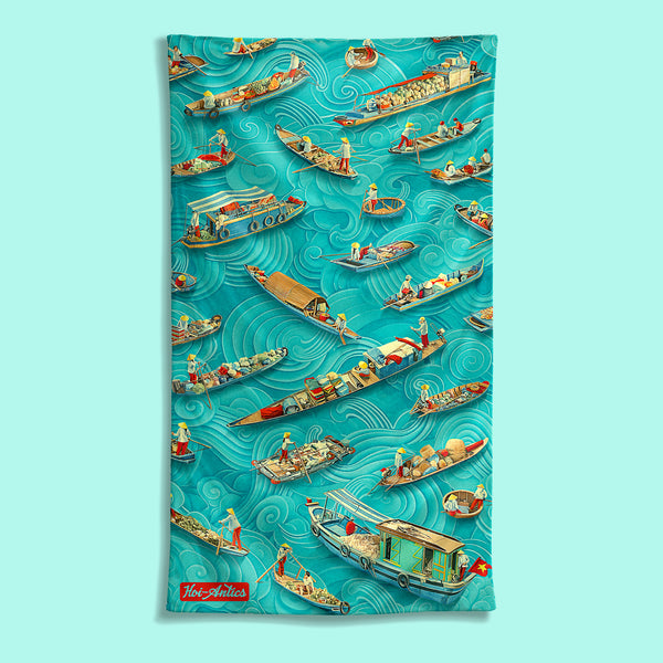 Front view of a large Mekong beach towel with a pattern of boats and people floating on asian inspired waves, featuring Hoi-Antics brand logo.