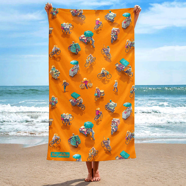 Front view of a large orange beach towel, held up at the beach, with a pattern of people on bicycles and motorcycles, featuring Hoi-Antics brand logo.