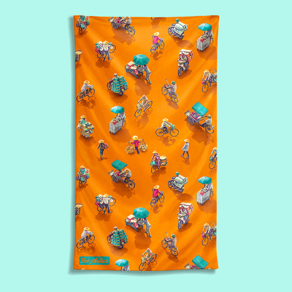 Front view of a large orange beach towel with a pattern of people on bicycles and motorcycles, featuring Hoi-Antics brand logo.