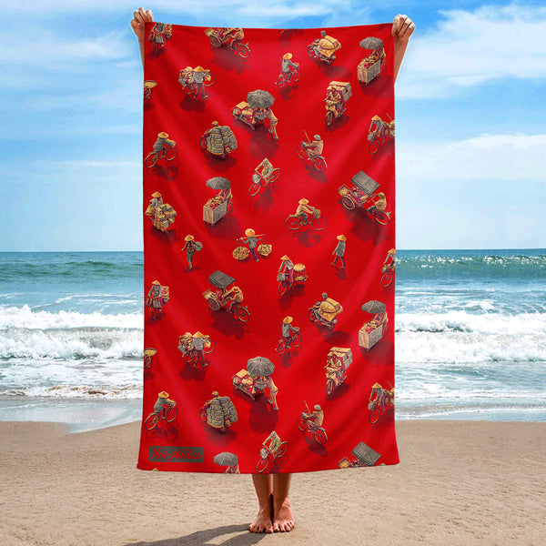 Front view of a large red beach towel, held up at the beach, with a pattern of people on bicycles and motorcycles, featuring Hoi-Antics brand logo.