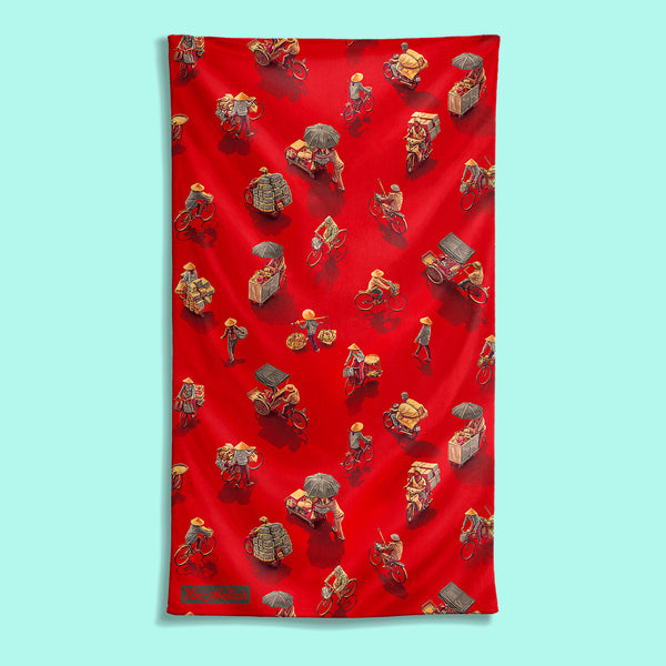 Front view of a large red beach towel with a pattern of people on bicycles and motorcycles, featuring Hoi-Antics brand logo.