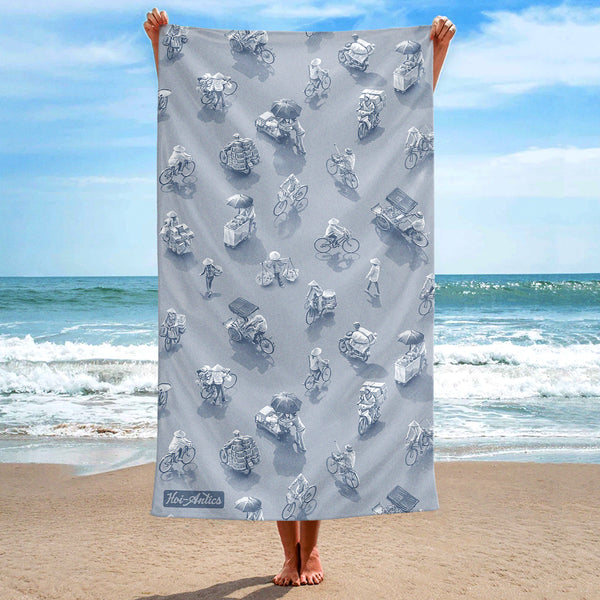 Front view of a large silver grey beach towel, held up at the beach, with a pattern of people on bicycles and motorcycles, featuring Hoi-Antics brand logo.