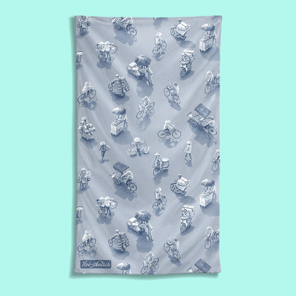 Front view of a large silver grey beach towel with a pattern of people on bicycles and motorcycles, featuring Hoi-Antics brand logo.
