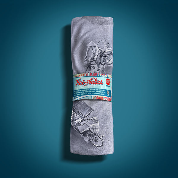 Front view of a rolled up large silver grey beach towel with a pattern of people on bicycles and motorcycles, featuring Hoi-Antics brand wrapper.