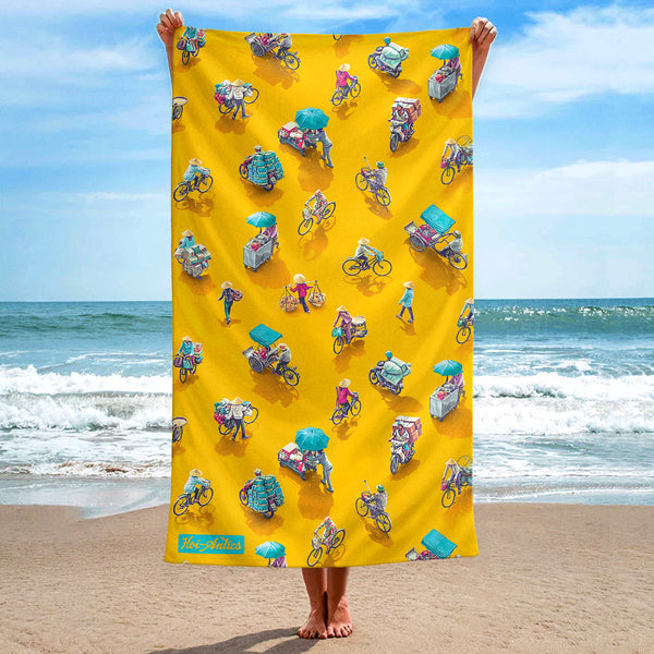 Front view of a large yellow beach towel, held up at the beach, with a pattern of people on bicycles and motorcycles, featuring Hoi-Antics brand logo.