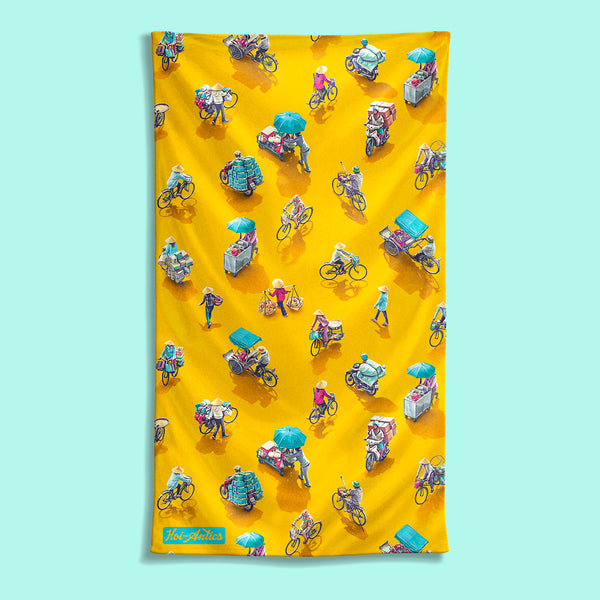 Front view of a large yellow beach towel with a pattern of people on bicycles and motorcycles, featuring Hoi-Antics brand logo.
