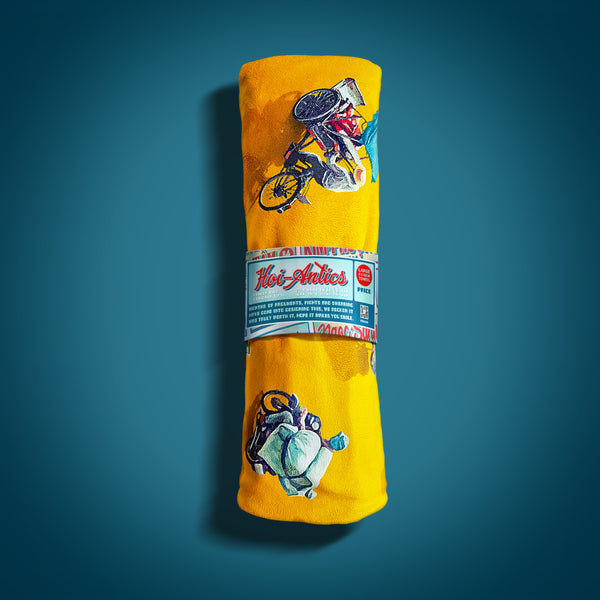 Front view of a rolled up large yellow beach towel with a pattern of people on bicycles and motorcycles, featuring Hoi-Antics brand wrapper.