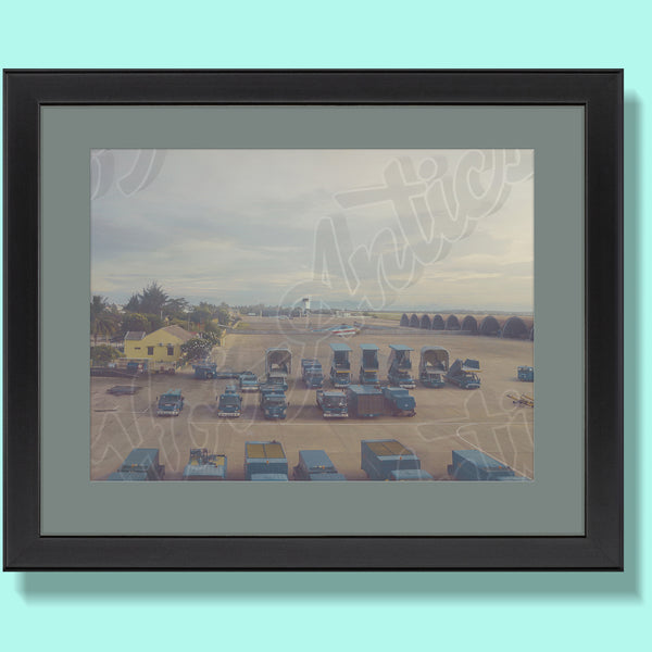 Front view of a Vietnamese airport setting, printed on fine art paper, framed and including helicopters, ground vehicles, buildings, hangers and a hazy afternoon sky.