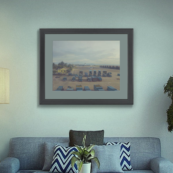 A Vietnamese airport setting, printed on fine art paper, framed and hanging on a lounge wall, including helicopters, ground vehicles, buildings, hangers and a hazy afternoon sky.